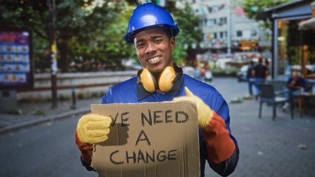 Construction worker holding cardboard sign reading we need a change, wearing hardhat and earmuffs, covers eye with gloved hand on street; workers rights hopeful.