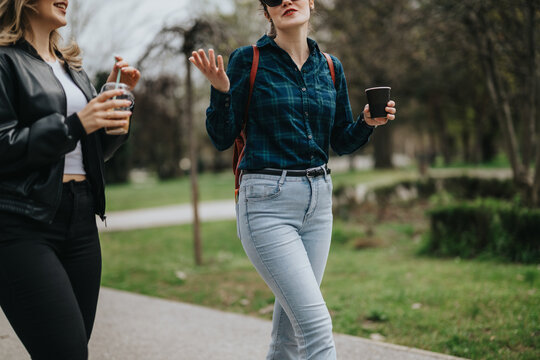 Two women stroll along a park path with coffee cups, casual outfits, and relaxed mood.