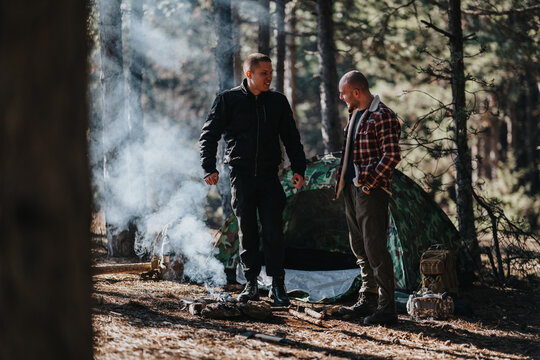 Two men stand near a campfire in a forest, with a camo tent in the background. They chat as smoke rises from the fire surrounded by trees.