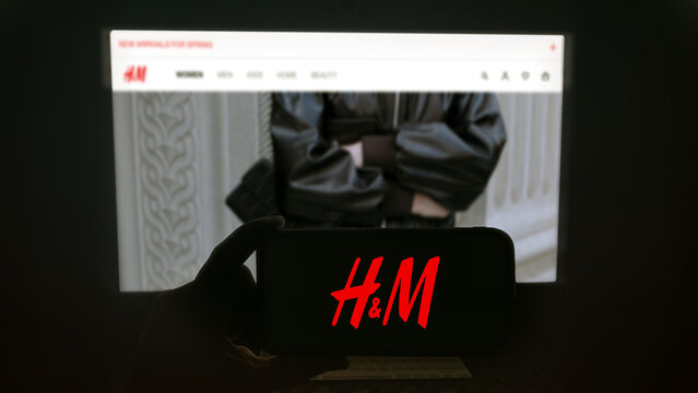 H & M app running on a modern mobile phone. The smartphone is being held in landscape mode, in the darkness. A PC monitor with the H & M website in the background.