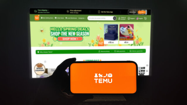 Temu app running on a modern mobile phone. The smartphone is being held in landscape mode, in the darkness. A PC monitor with the Temu website in the background.