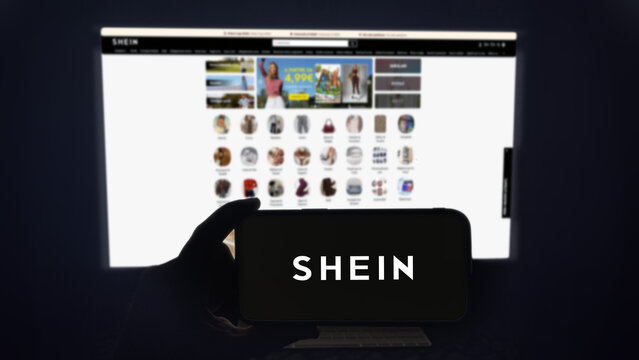 Shein app running on a modern mobile phone. The smartphone is being held in landscape mode, in the darkness. A PC monitor with the Shein website in the background.