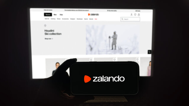 Zalando app running on a modern mobile phone. The smartphone is being held in landscape mode, in the darkness. A PC monitor with the Zalando website in the background.