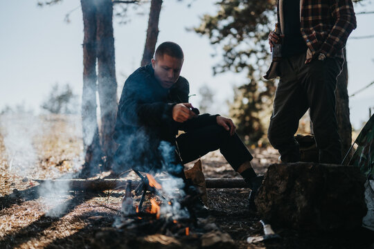 Two men gather around a small campfire in a woodland setting; one sits close to the flames.