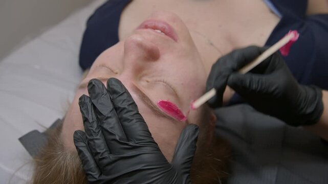 Beautician spreads warm green wax on eyebrows for shape correction and removes excess hair with strip. Procedure after permanent makeup.Woman in salon,Precise PMU preparation and clean contour result.