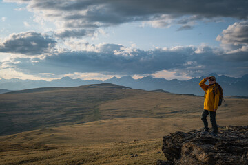 Fototapeta premium Female hiker in yellow jacket standing on edge of rocky cliff looking at vast mountain plateau under cloudy sky during sunset travel adventure. 