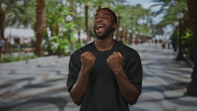 Young african american man with clenched fists and visible braces smiling on busy seaside street promenade with palm trees; triumph.