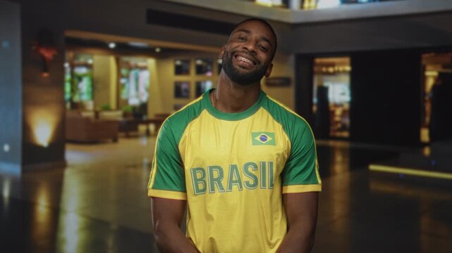 Man smiling, standing in a hotel building lobby wearing a yellow and green brazil jersey with flag patch, facing camera; pride.