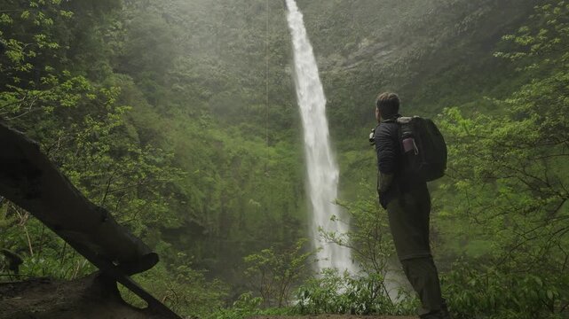One person rappels beside waterfall as another walks closer from the forest floor to watch