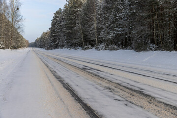 Obraz premium a paved road covered with ice for cars through a winter forest in the snow , a narrow track covered with snow from cars in the forest in the winter season, different types of trees grow