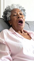 Elderly Black woman yawning on gray couch, wearing a pink pajamas top, against a plain white wall backdrop