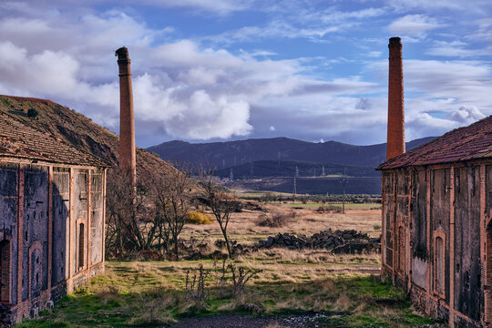 View of weathered brick structures frame a sprawling field under a textured sky, leading to distant mountains and wind turbines, Puertollano, Castile-La Mancha, Spain.