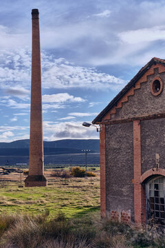View of a towering brick chimney piercing the cloudy sky beside a decaying building, a stark contrast against the grassy field, Puertollano, Castile-La Mancha, Spain.
