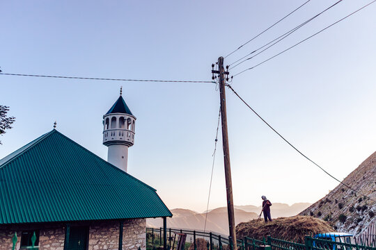 View of a man working on a haystack next to a building with a green roof and a minaret rising against the mountain backdrop, Lukomir, Federation of Bosnia and Herzegovina, Bosnia and Herzegovina.