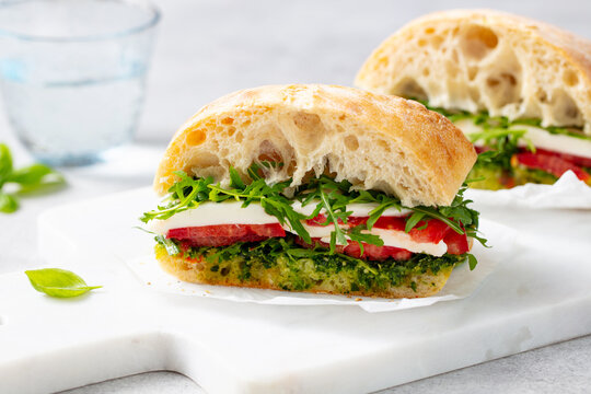 Ciabatta sandwich with mozzarella, tomatoes, pesto sauce and fresh basil on marble serving board. Light grey background. Close up.