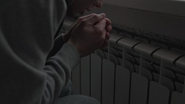 Cold apartment lifestyle, man warming hands near radiator, freezing at home during winter heating problems.