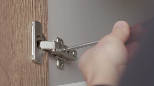 Hands tighten cabinet hinge screw with screwdriver on wooden door. Concept of cabinet hinge installation and home repair maintenance.
