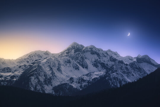 View of snow-capped peaks rise majestically under a gradient sky where golden light meets the deep blue of twilight, watched over by a crescent moon, Trentino-South Tyrol, Italy.