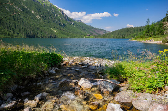View of a serene lake reflecting the surrounding verdant mountains under a bright sky, with a rocky stream flowing into it, Morskie Oko, Wysokie Tatry, Wojewodztwo malopolskie, Poland.