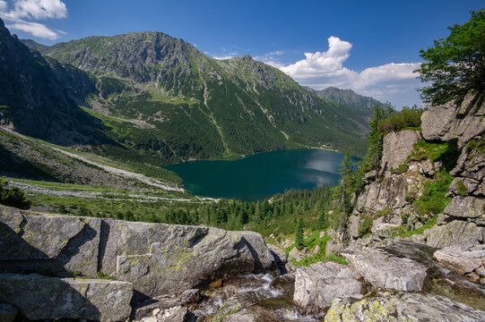 View of a pristine lake nestled amidst towering mountains, with jagged rocks framing the foreground in a scene of natural majesty, Morskie Oko, Wysokie Tatry, Poland.