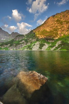 View of serene waters reflecting the towering mountains under a blue sky dotted with fluffy clouds, a boulder sits in the foreground, Morskie Oko, Wysokie Tatry, Poland.