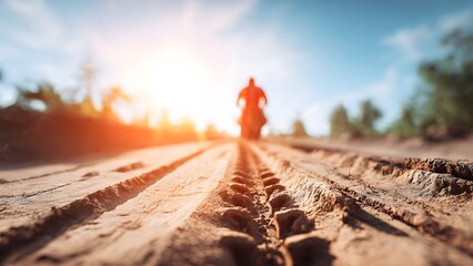 Deep tire tracks carved in a dusty dirt road lead towards a blurred motorcyclist silhouette against a bright sun evoking adventure and offroad journey freedom.