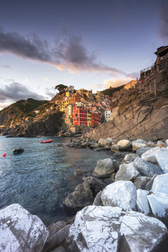 View of colorful buildings clinging to rugged cliffs, with azure waters lapping against the rocky shore under a vibrant sky, Riomaggiore, Liguria, Italy.