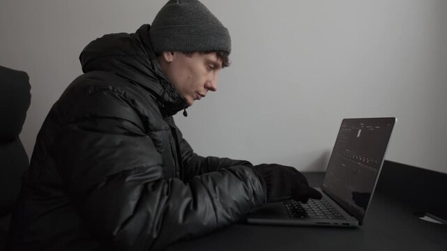 Human adaptation to harsh living conditions, man working on laptop in winter jacket, hat and gloves at home. Cold home office