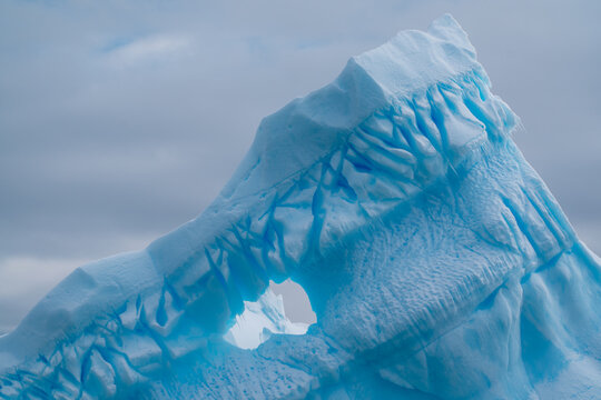 View of a colossal iceberg, sculpted by wind and water, displaying a fascinating hole against the backdrop of a cloudy sky, Portal Point, Graham Land, Antarctica.