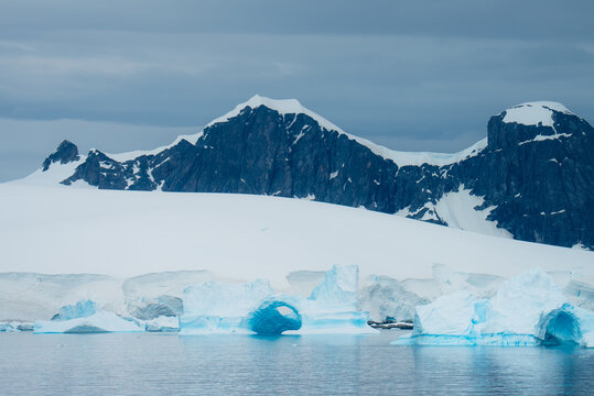 View of shimmering cerulean icebergs drift in the frigid waters beneath snow-capped mountains under a steel-grey sky, Portal Point, Antarctica.