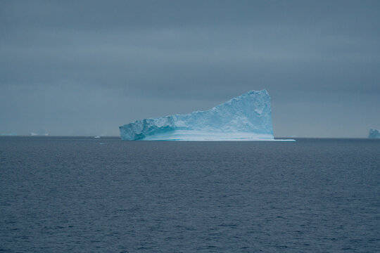 View of a massive, angular iceberg, a frozen sculpture of nature, drifts serenely in the dark, frigid waters under a heavy, overcast sky, Portal Point, Antarctica.