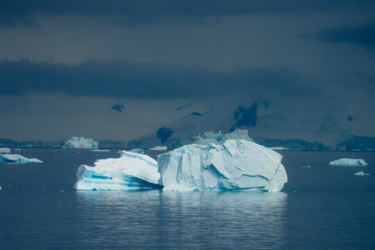 View of a pristine iceberg floats majestically on dark waters under a clouded sky, framed by distant, snow-capped mountains, Portal Point, Antarctica.
