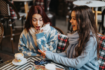 Fototapeta premium Two young women in sweaters sit at a patterned table in a cafe, one surprised and the other smiling. They enjoy coffee, conversation, and a friendly moment between friends.