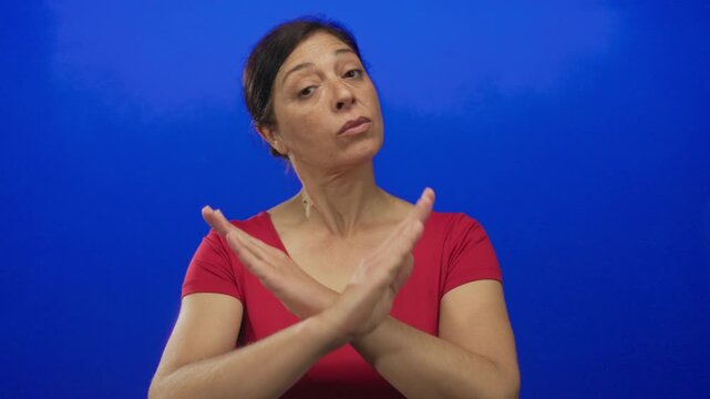 Hispanic middle age woman crossing forearms in studio against blue backdrop wearing red top; rejection.