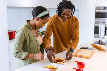 African American couple preparing flatbread on white island, spreading sauce and steadying dish © wavebreak3