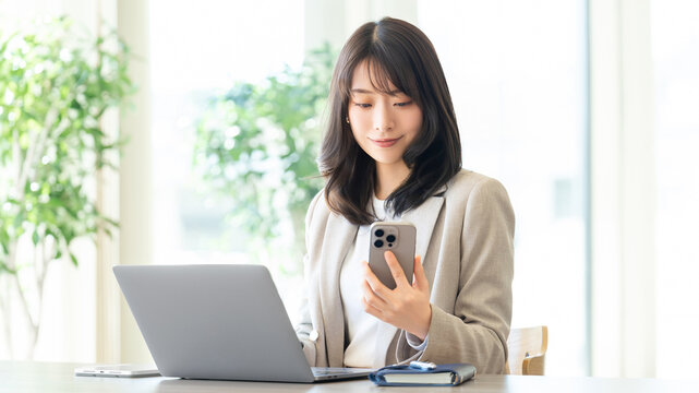 ノートパソコンを前にスマートフォンを確認する女性ビジネスパーソン, Businesswoman checking smartphone in front of laptop