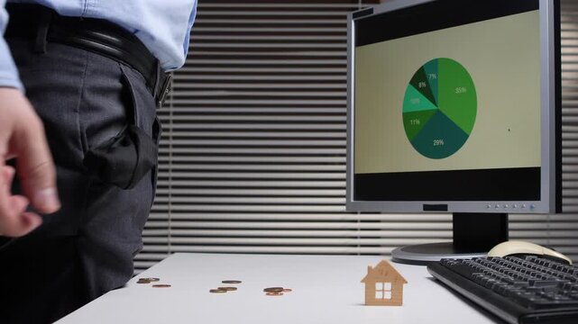 Man pulls out empty pocket at desk with coins and wooden house model beside a computer showing a pie chart, symbolizing financial strain, mortgage pressure and budgeting challenges