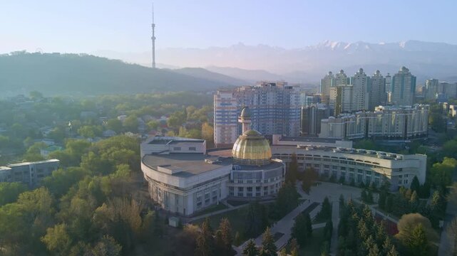 Cinematic aerial drone flight over Almaty city skyline. Panoramic view of modern skyscrapers and historical buildings with snow-capped Tian Shan mountains in Kazakhstan.