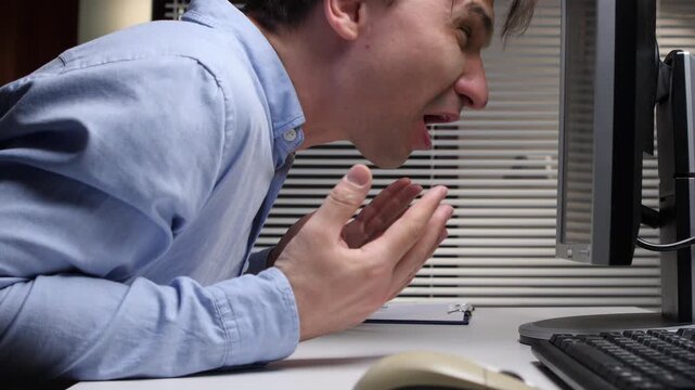 Man in a light blue shirt expressing extreme anger and frustration while screaming at a computer monitor in an office setting, possibly experiencing technical problems or work related stress