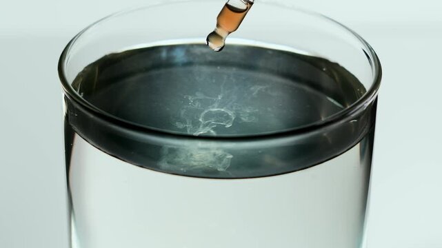 Macro shot of a glass pipette releasing an amber serum droplet into clear water. In slow motion, the liquid spreads in soft cloudy diffusion inside the glass on a light minimal background.