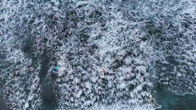 Aerial view of surfers riding waves in the ocean. Multiple unrecognizable people on surfboards navigating through the water, with dynamic wave patterns and foamy surf visible in the background