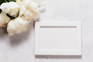 Horizontal white wooden picture frame mockup on a grey concrete background next to fresh white peony flowers. Flat lay composition provides empty space for artwork, photography, or wedding text.