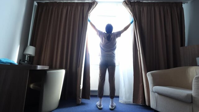 A maid in a hotel room, wearing gloves, opens the curtains to let in natural light.