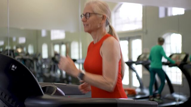 Mature woman runs on treadmill in gym while engaged in her daily exercise routine alongside other gym members in the afternoon.