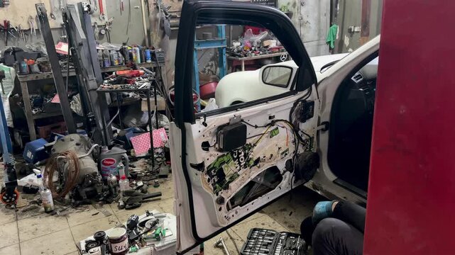 Auto mechanic working on a car door repair in a professional garage. Close-up of disassembled white car door with internal mechanisms, wiring, and tools visible in a workshop background.