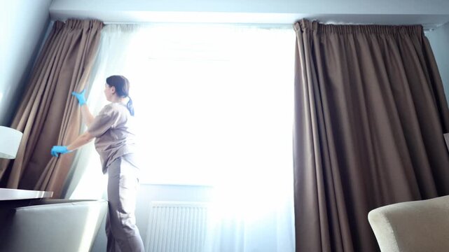 A maid in a hotel room, wearing gloves, opens the curtains to let in natural light.