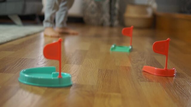  A dynamic shot of a child playing with various mini golf flags and holes spread across the room, showing movement, playfulness, and active indoor recreation.