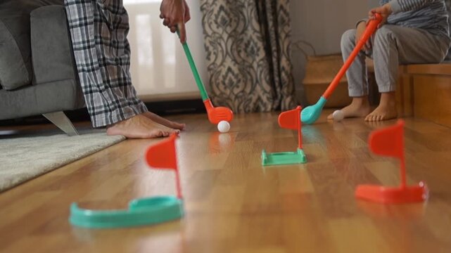A wide shot of a living room mini-golf circuit where the child mischievously hits the ball right before the father&rsquo;s swing.