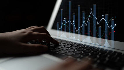 Hands typing on a laptop keyboard display visual analytics and financial trends in a dark...
