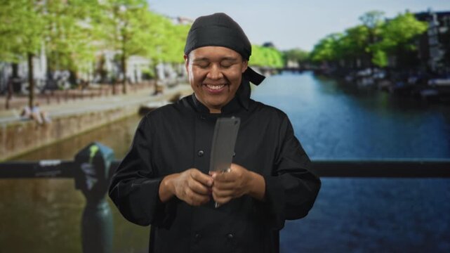 Man chef in black uniform points fingers to smile while holding kitchen knife on a canal street in amsterdam; joyful playfulness hospitality.